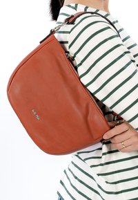 Brūns ādas rokassomiņa ar izliekta formu, sudraba rāvējslēdzēju un regulējamu siksnu, turēta pret svītrainu krekls. Gluda tekstūra.