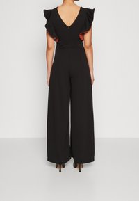 Mujer vestida con un mono negro de pierna ancha, con mangas cortas con volantes y un escote en V profundo, de pie contra un fondo claro y liso.
