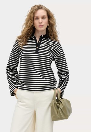 Frau mit lockigem Haar, die ein schwarz-weiß gestreiftes Langarmshirt und cremefarbene weite Hosen trägt und eine olivgrüne Handtasche hält.