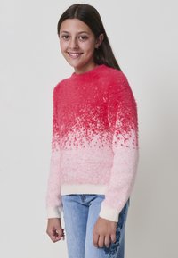 Fille souriante, portant un pull dégradé rouge à rose duveteux et un jean bleu clair avec broderies florales, se tenant contre un fond blanc uni.