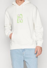 Sweat à capuche blanc avec une poche avant, présentant un motif graphique coloré de lettres et de formes. Fabriqué en tissu doux avec une capuche à cordon.