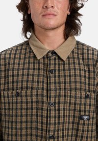 Camisa de cuadros marrón y negra, con un collar de tela de pana beige, que presenta botones negros y un pequeño logo en el bolsillo del pecho.