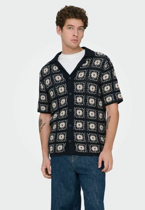 Ung mand med krøllet hår iført en sort og hvid mønstret kortærmet cardigan med knapper over en hvid skjorte og blå jeans.