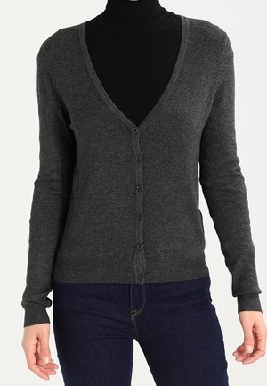 Femme portant un cardigan gris foncé boutonné sur un col roulé noir et un jean bleu foncé, debout devant un fond uni.