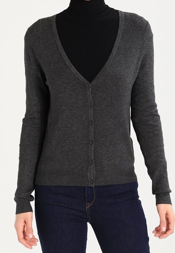 Femme portant un cardigan gris foncé boutonné sur un col roulé noir et un jean bleu foncé, debout devant un fond uni.
