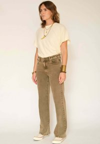 T-shirt beige clair, jean flare vert olive avec cinq poches. Porté avec des baskets blanches et des bijoux superposés couleur or. Texture lisse.