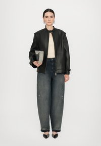 Giacca di pelle nera con colletto alto, corpetto attillato e zip. Jeans blu a gamba larga, perline sulla pochette e scarpe nere a punta.
