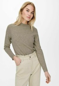 Maglione lavorato a maglia verde oliva con un collo alto e maniche lunghe, indossato con pantaloni beige a vita alta dotati di un elastico in vita.