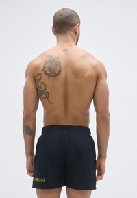 Sötétkék fürdőnadrág, gumis derékrésszel és sárga logóval. A személy hátán látható egy kígyó és szív mintájú tetoválás.