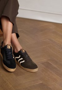 Black and brown sneakers featuring a leopard print tongue, white stripes, and gum soles. Material is a mix of suede and canvas.