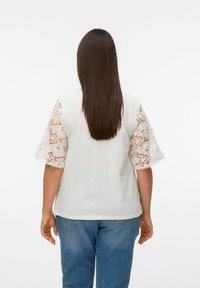 Witte T-shirt met kant korte mouwen, voorzien van bloemmotieven. Gecombineerd met blauwe jeans, waardoor er een casual outfitoptie ontstaat.