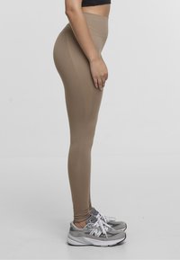 Leggings de talle alto en color beige, hechos de tela elástica, con una textura suave y detalles en la costura lateral, combinados con zapatillas deportivas grises.