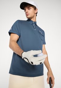 Mørkeblå kortærmet polo t-shirt med knaplukning og brystlomme. Hvid handske der holder en golfbold, beige shorts og en hvid kasket.