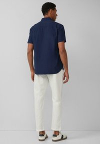 Marineblaues Kurzarmhemd, weiße taillierte Hose und niedrige Sneaker mit schwarz-weißen Akzenten. Glatter Stoff und klare Linien.