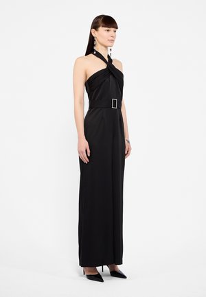 Zwart halter jumpsuit met gedraaide buste, brede riem met een rechthoekige gesp en tapered broekspijpen, gecombineerd met zwarte puntige hakken.