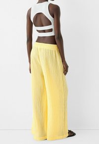 Vit ärmlös topp med skuren rygg, matchad med livfullt gula plisserade vida byxor. Observera: mjuk textur, elastisk midjeband.