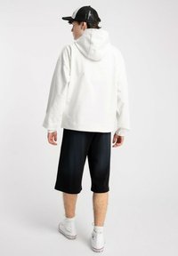 Młoda osoba ubrana w białą bluzę z kapturem, czapkę z czarnej siateczki, czarne szorty do kolan i białe trampki za kostkę, stojąca tyłem na jednolitym tle.