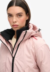 Jasnoróżowa wodoodporna kurtka z czarnym wewnętrznym kołnierzem, pełnym zamkiem błyskawicznym i regulowanym kapturem. Posiada czerwone logo na ramieniu.