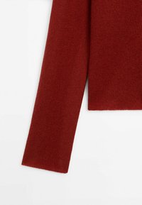 Détail d'un pull rouge en tricot, avec une manche longue et droite et un ourlet net. Texture douce avec une finition unie, sans motifs ni embellissements.