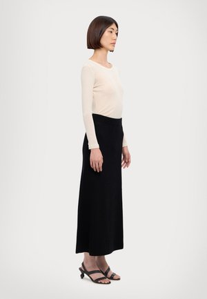 Jupe longue noire à côtes associée à un haut léger beige à manches longues. La personne porte des sandales noires à lanières. Le design est ajusté et simple.
