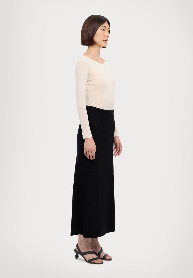 Lang sort ribbet nederdel parret med en langærmet lys beige top. Personen har sorte stroppesandaler på. Designet er tætsiddende og enkelt.