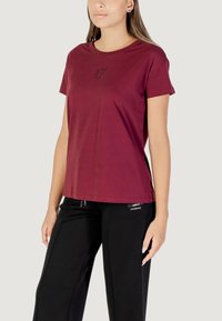 T-shirt in cotone bordeaux con maniche corte, scollatura rotonda e un sottile dettaglio di logo con paillettes sul davanti. Abbinato a pantaloni sportivi neri.