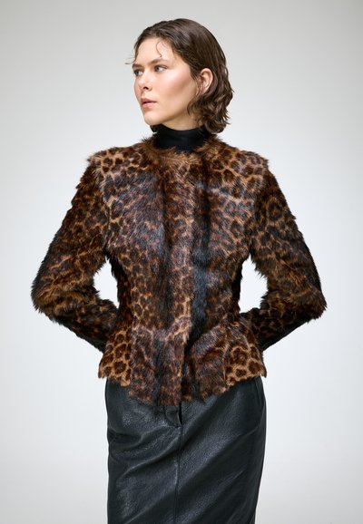 Femme aux cheveux courts et bouclés portant une veste en fourrure à motif léopard et une jupe en cuir noire, regardant vers la gauche avec les mains sur les hanches.