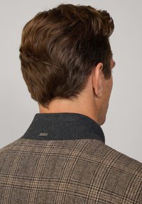 Saco deportivo a cuadros con un patrón marrón y gris, cuello negro en contraste y acento de logo metálico en el cuello. Tela texturizada.