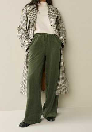 Person, die einen beigen Rollkragenpullover, einen karierten langen Mantel, weite grüne Cordhose und schwarze Loafers trägt, steht vor einer schlichten Wand.