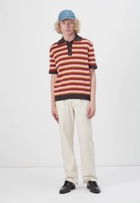 Strikket polo skjorte med sikksakkmønster i rødt, oransje og gult, svart krage. Kombinert med lysebeige bukser og svarte sko, iført en blå caps.