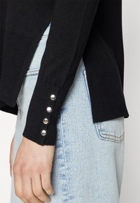 Μαύρο πλεκτό πουλόβερ με πλευρική σχισμή, ραβδωτή υφή και ασημένια κουμπιά στα μανίκια. Συνδυάζεται με ανοιχτόχρωμο μπλε τζιν.