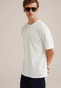 Camiseta corta de manga blanca con textura y cuello redondo, que presenta un pequeño logotipo cerca del dobladillo, combinada con pantalones oscuros y gafas de sol.