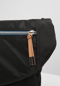 Borsa di stoffa nera con una tasca con cerniera dotata di cerniera blu e un manico in pelle marrone, superficie testurizzata e design a pattina pieghevole.