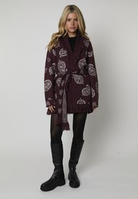 Cárdigan de punto burdeos con patrones de paisley blancos, bordes con flecos y un cinturón a juego, llevado sobre una blusa negra y combinado con botas negras hasta la rodilla.