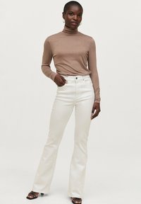 Lys beige turtleneck-top kombineret med højtaljede, flagrende hvide jeans. Model iført sorte strop sandaler med minimal smykkeunivers.