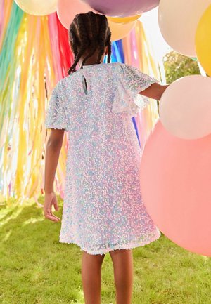 Robe en sequins irisés avec manches courtes évasées, ouverture en forme de goutte dans le dos et ourlet festonné, sur fond de ballons colorés.