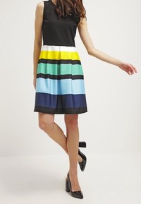 Czarny top bez rękawów połączony z wielokolorową spódnicą w paski w kolorze żółtym, zielonym, niebieskim i czarnym; gładka struktura; w zestawieniu z czarnymi butami na obcasie.