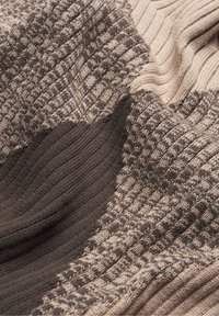 Lähellä otettu kuva pehmeästä neulotusta kankaasta, jossa on röyhelö- ja tekstuurointikuvioita beige- ja ruskeasävyissä.