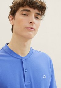 Modrá polokošile s texturovaným povrchem, vybavená dvouknoflíkovou légou a malým bílým logem na levé straně.