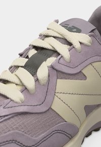 Κλείσιμο σε ένα μοβ αθλητικό παπούτσι με κρέμα κορδόνια και μπεζ ραφές επικαλύψεις που εμφανίζουν υφασμάτινη υφή και επενδεδυμένη γλώσσα.