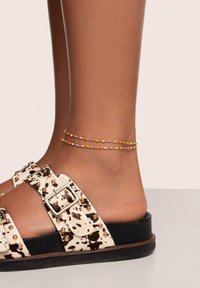 Schwarz-weiße gemusterte Sandale mit goldenen Nieten, ausgestattet mit zwei zarten Knöchelarmbändern in Gelb und mehrfarbigen Perlen.
