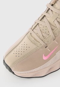 Sneaker bege da Nike com logótipo swoosh rosa, área do dedo perfurada, atacadores trançados e sola de borracha sobre um fundo claro.