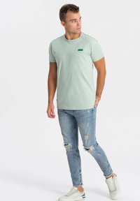 Světle zelené bavlněné tričko s kulatým výstřihem, zkombinované s odrbanými světle modrými džíny a bílými teniskami s šedým a zeleným akcentem.