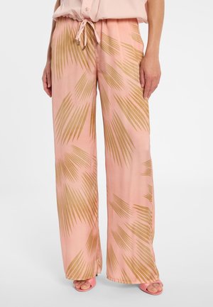 Frau trägt rosa weite Hose mit braunen abstrakten Palmblattmustern und passende rosa offene Schuhe mit Absatz.