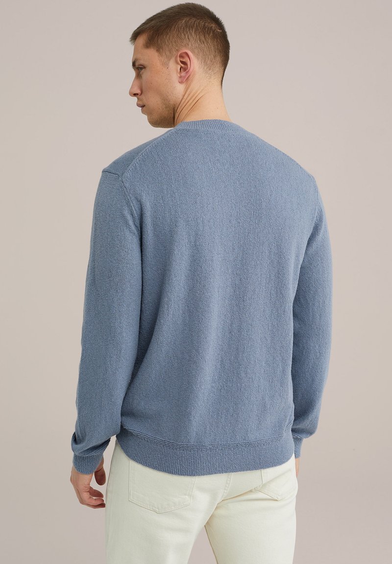 Gaiši zils adīts džemperis ar struktūrētu virsmu, apaļa kakla izgriezums, ribota piedurknes un apakša, valkāts ar gaišas krāsas biksēm, skatīts no aizmugures.