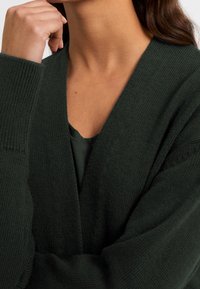 Mörkgrön kofta i stickad tyg, med djup V-halsringning och ribbade ärmar. Mjuk textur med avslappnad passform.