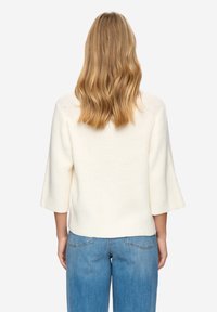 Krémový pletený svetr se tříčtvrtečními zvonovými rukávy, žebrovanou strukturou a kulatým výstřihem, v kombinaci s světle modrými džínami.