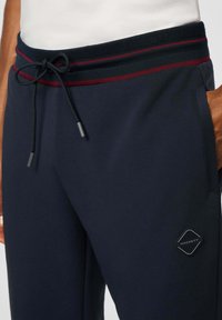 Marineblauwe sweatpants hebben een gestructureerde tailleband met rode en groene strepen, een trekkoord, een zijzak en een rubberen logo-embleem.