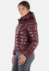 Giacca a vento bordeaux con superficie lucida, trapuntatura verticale, cappuccio integrato e tasche con zip. Presenta polsini elastici e dettaglio con logo.