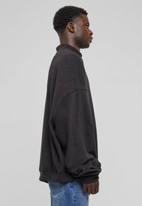 Overdimensjonert svart sweatshirt i myk stoff, med høy krage, senket skuldersømmer og ribbede mansjetter. Kombinert med lyseblå jeans.